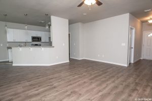 Empty room with wood flooring and white walls next to a kitchen area.